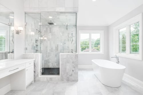 Bright luxury bathroom remodel featuring a marble walk in shower with frameless glass, freestanding soaking tub, white vanity, and large windows with natural light.