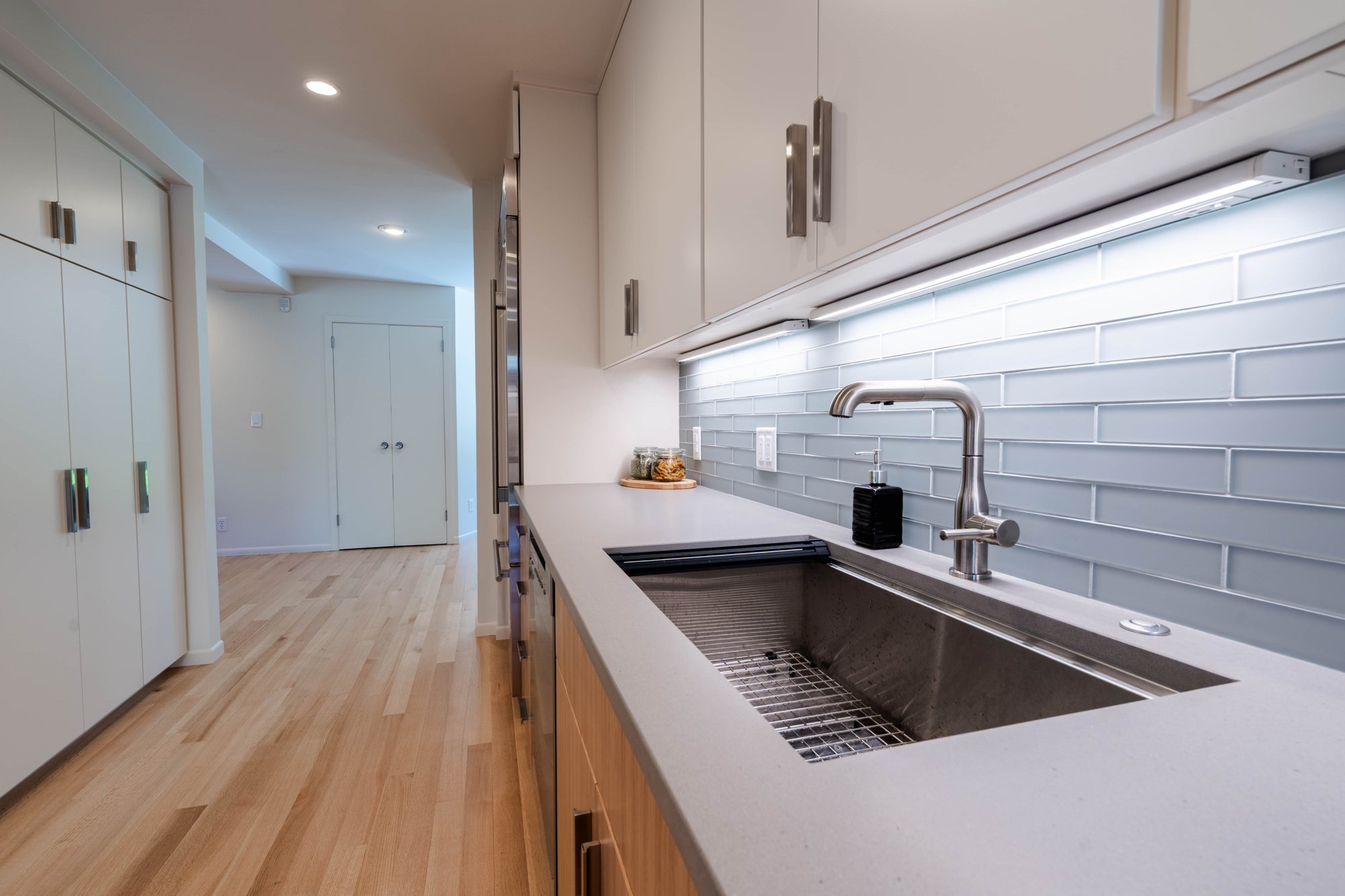 Kitchen sink with stainless steel faucet, gray tile backsplash, under-cabinet lighting, and white countertops.