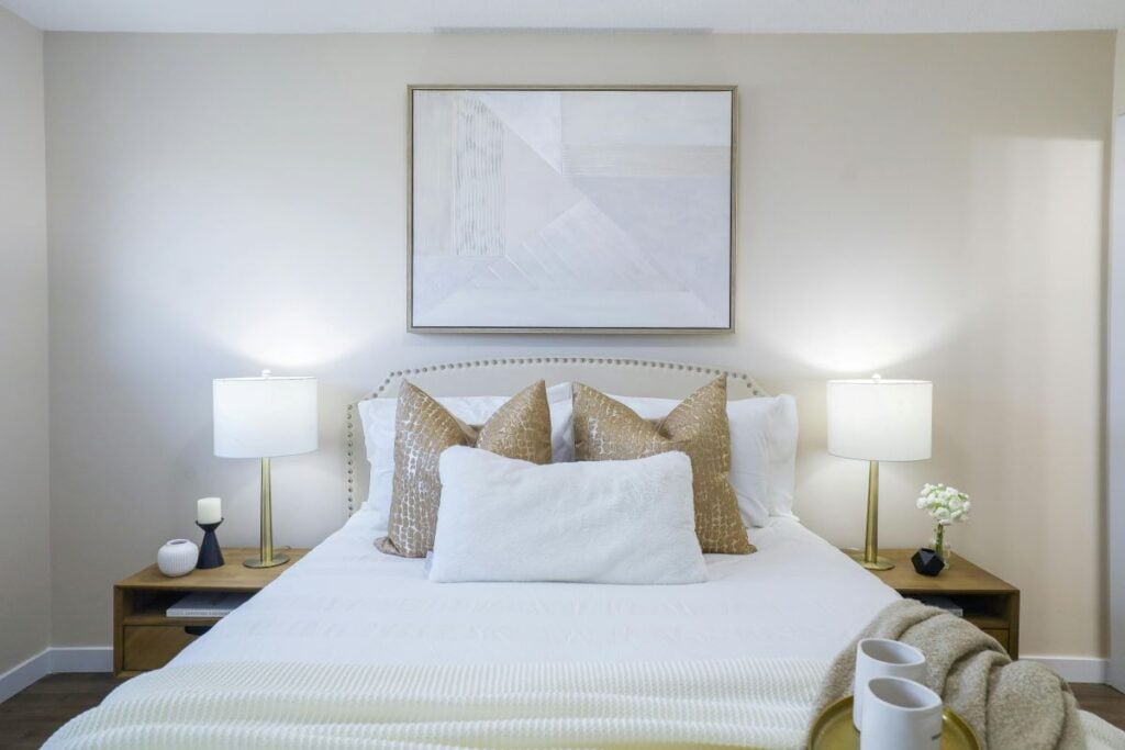 Bright, neutral-toned bedroom with layered white bedding, gold accent pillows, matching bedside lamps, and wall art.