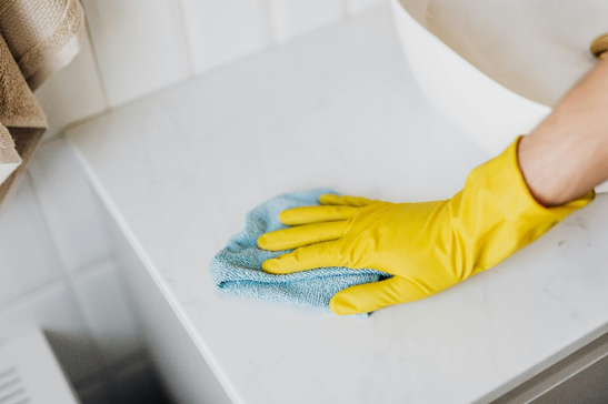 Person wearing yellow cleaning gloves wiping down a bathroom countertop with a cloth