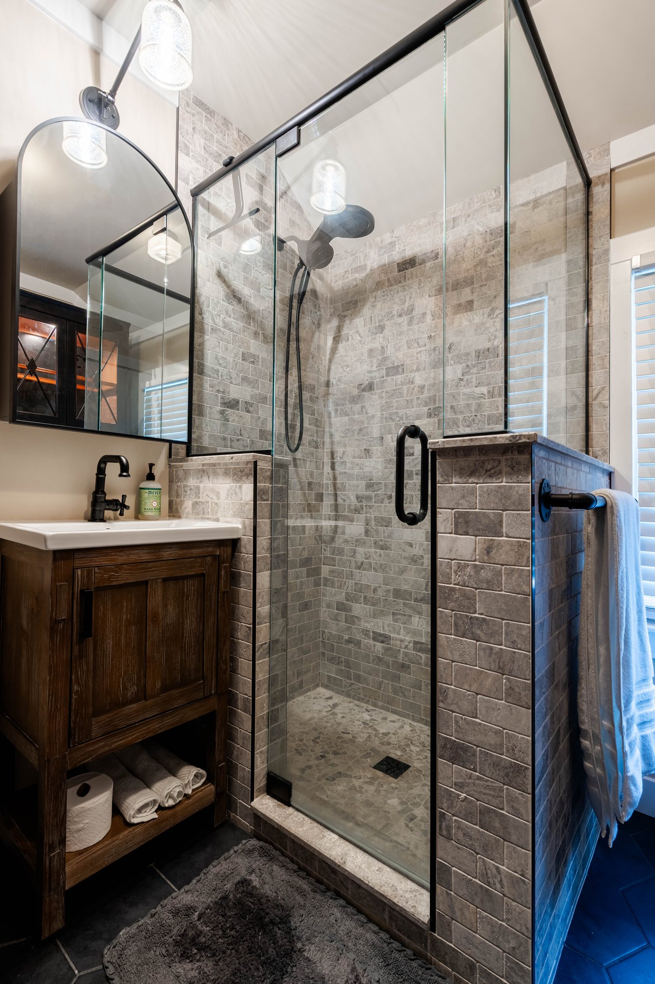 Natural stone tiled shower remodeled by Homecare Remodeling with a black-framed glass enclosure, pebble tile floor, and matte black fixtures.