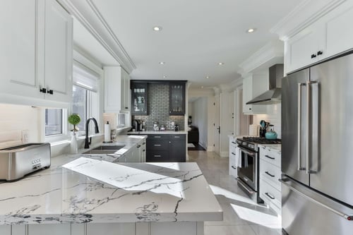 Remodeled kitchen featuring white shaker cabinets, quartz countertops with bold veining, a large peninsula, black accents, and stainless steel appliances.
