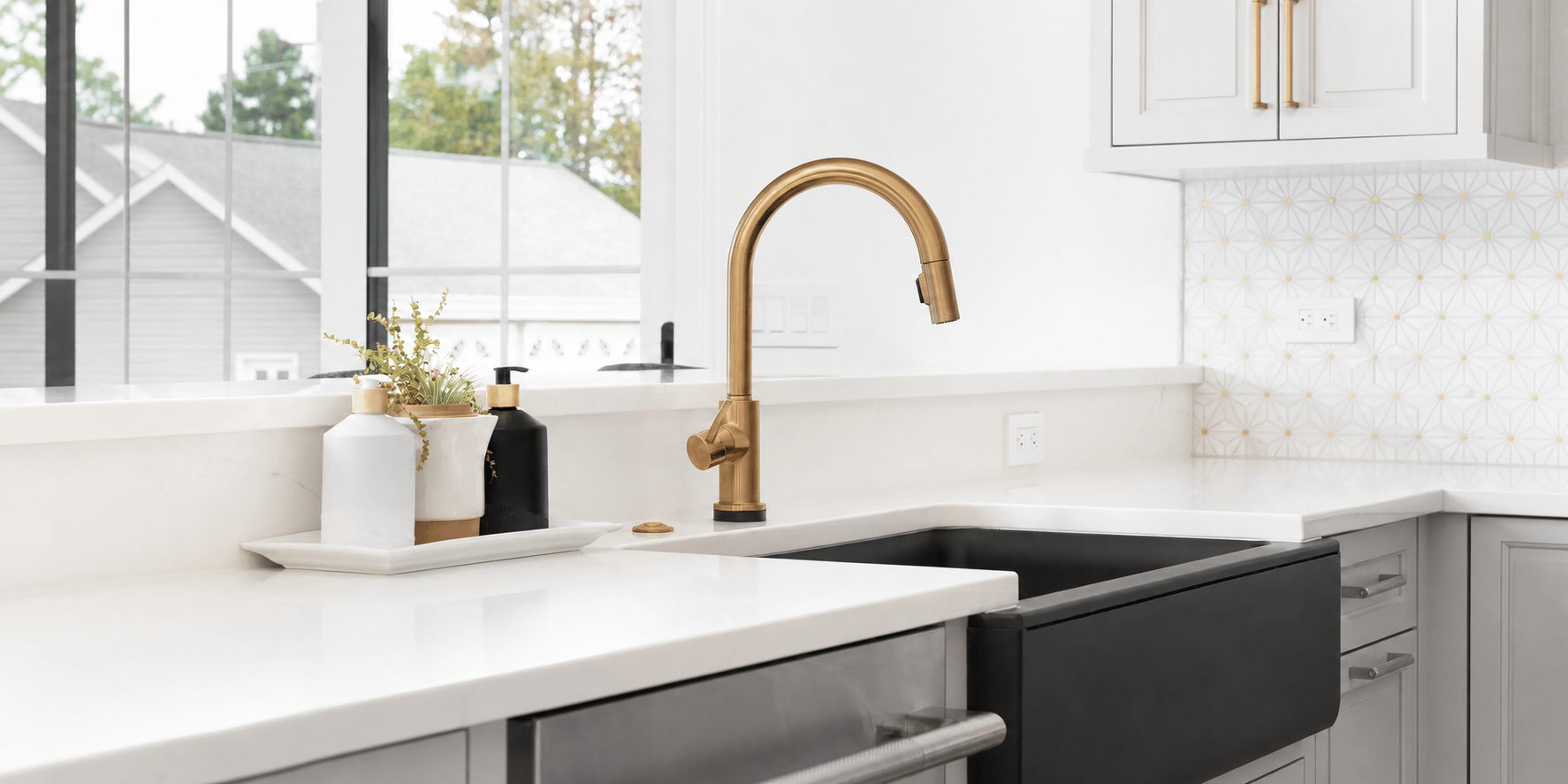 Modern kitchen sink with a black farmhouse basin, brushed brass faucet, white countertops, and white cabinetry.