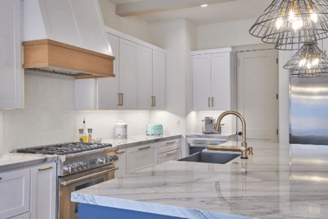 White kitchen with a marble island, wood-trimmed range hood, stainless steel appliances, and wire pendant lights.