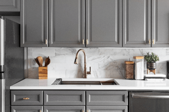 Gray kitchen cabinets with brass hardware, a marble tile backsplash, white countertop, and undermount sink.