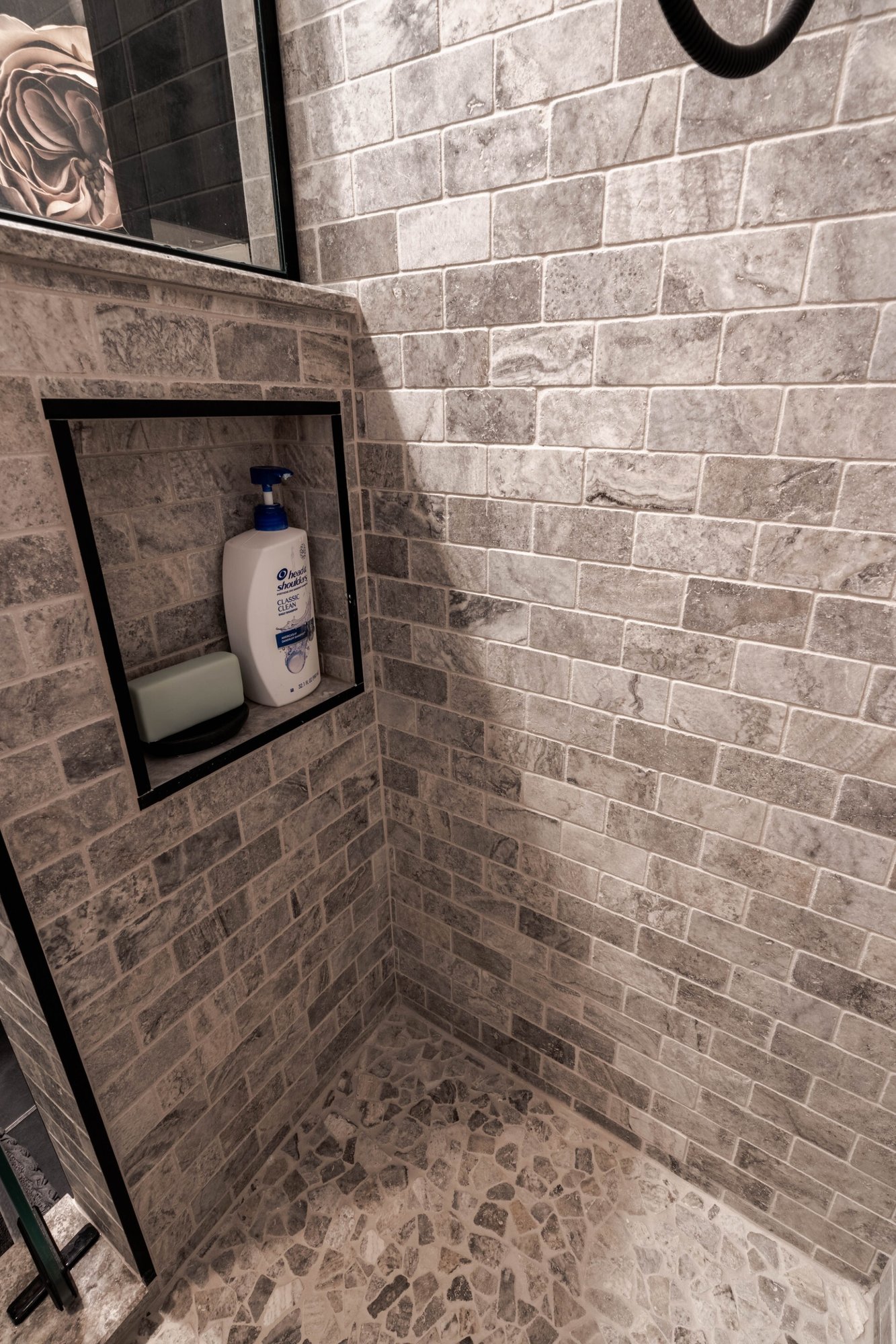 Natural stone tiled shower remodeled by Homecare Remodeling featuring a recessed niche with black trim and a pebble tile floor.