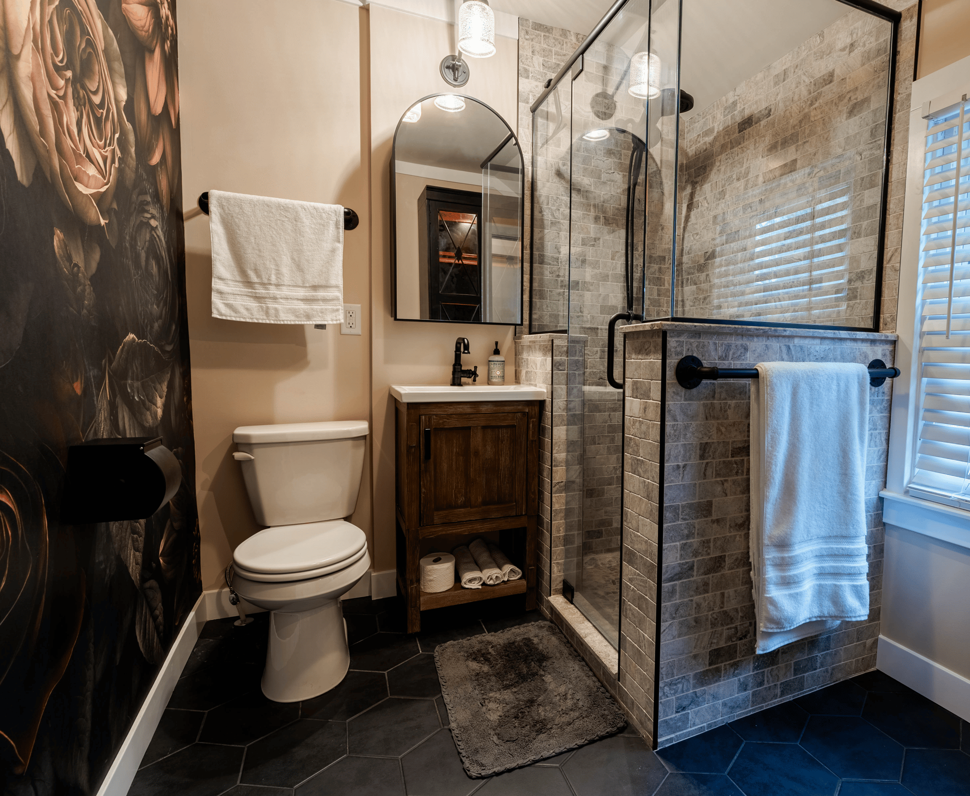 Minneapolis bathroom remodeled by Homecare Remodeling featuring a natural stone tiled shower, black-framed glass enclosure, wood vanity, and floral accent wall.