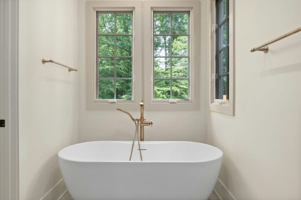 Freestanding white bathtub with gold fixtures beneath tall windows.