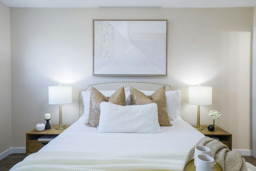 Bright, neutral-toned bedroom with layered white bedding, gold accent pillows, matching bedside lamps, and wall art.