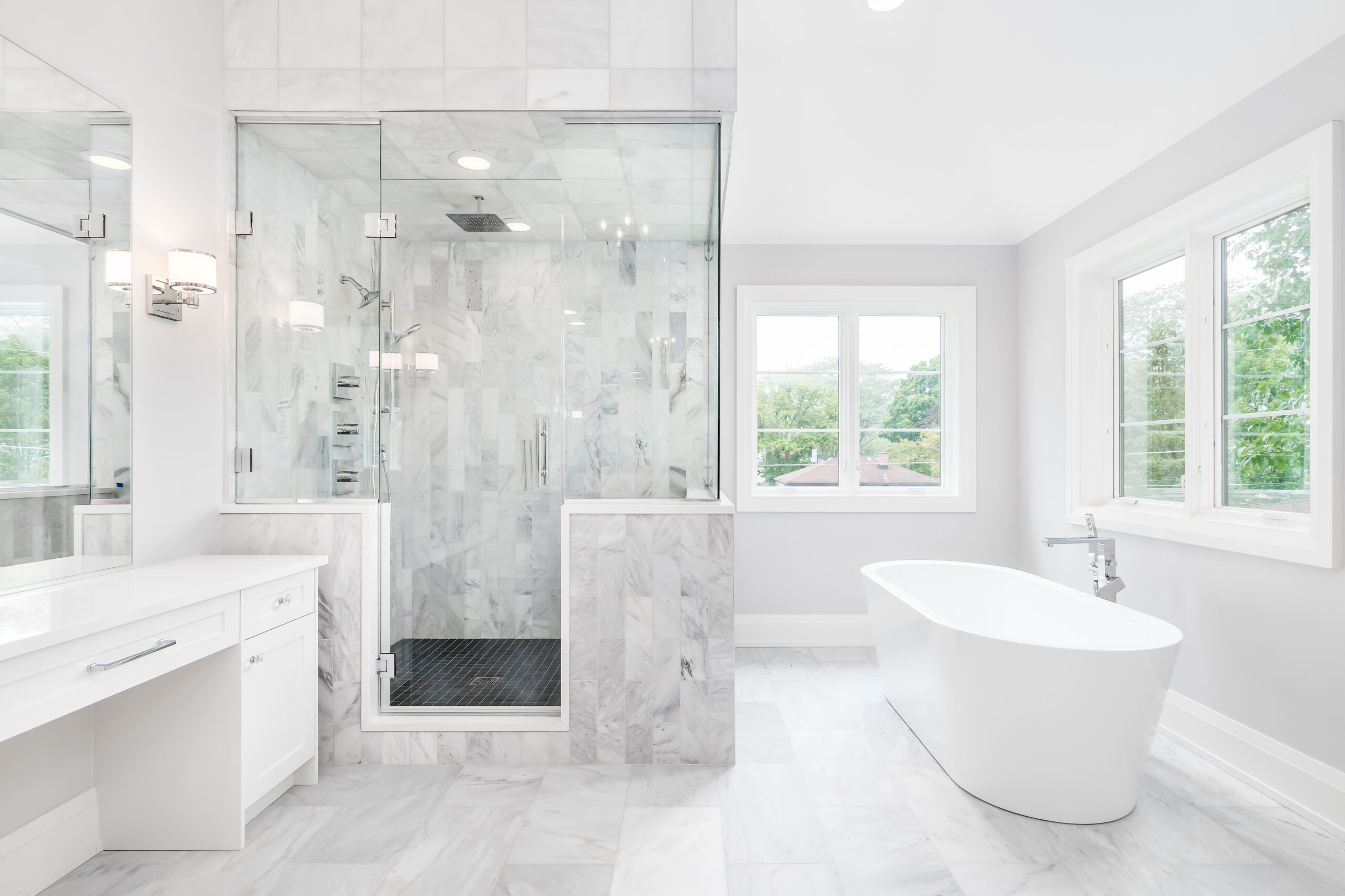 Bright luxury bathroom remodel featuring a marble walk in shower with frameless glass, freestanding soaking tub, white vanity, and large windows with natural light.